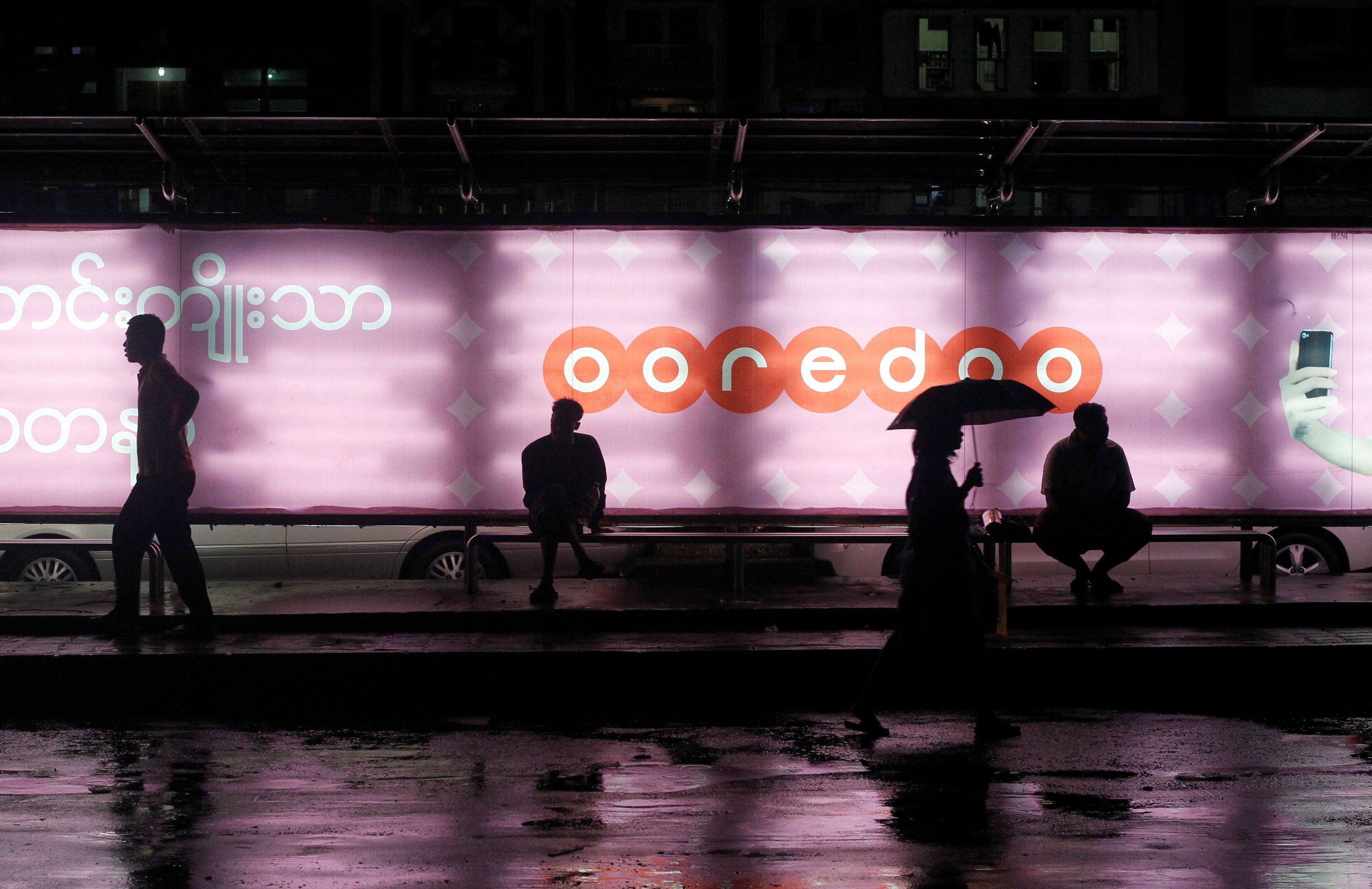 People sit at a bus station with an Ooredoo advertisement as they wait for a bus in Yangon