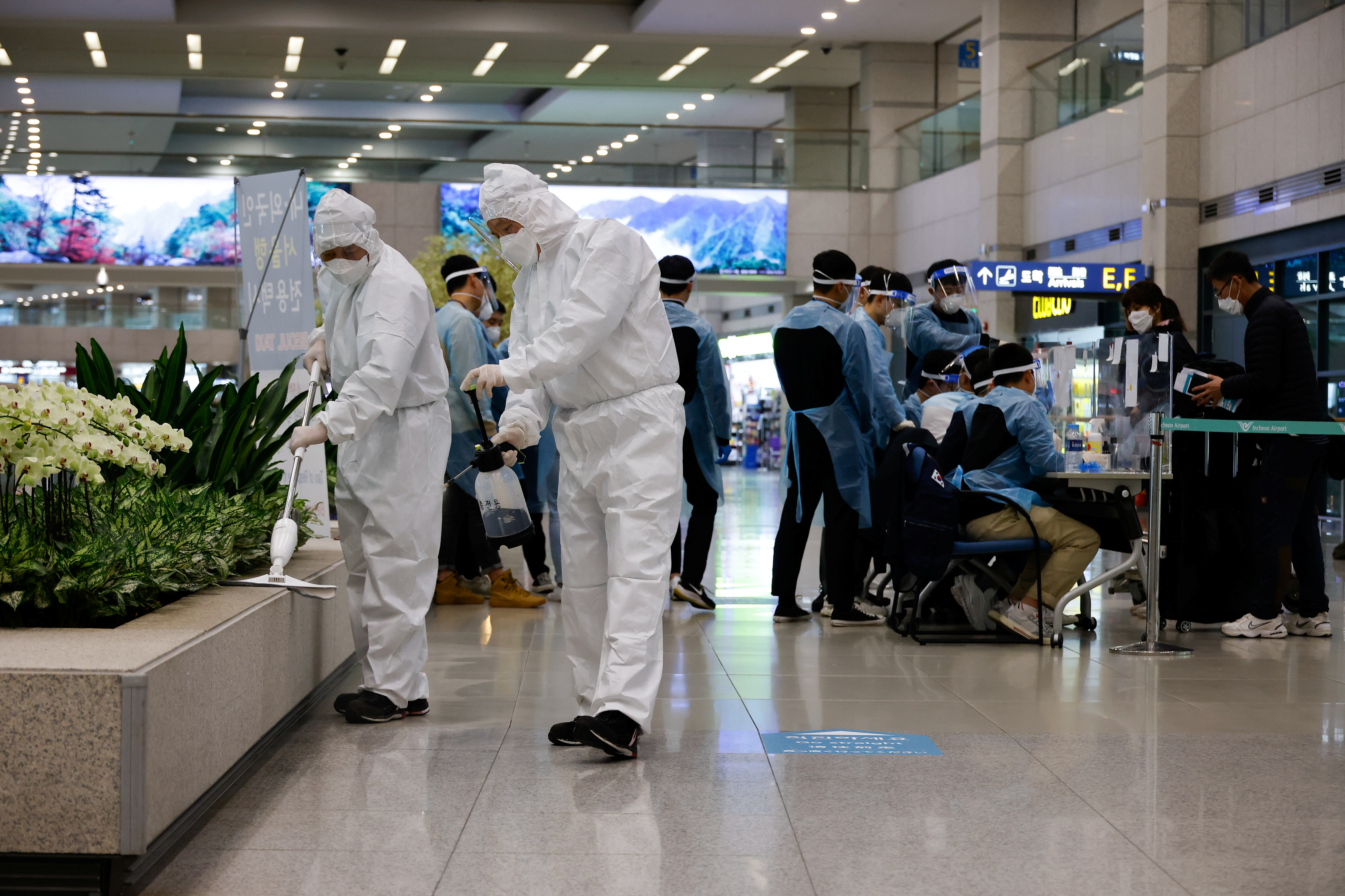 Workers wearing protective gear disinfect an arrival gate as other workers check passengers from overseas upon their arrival at the Incheon International Airport, in Incheon