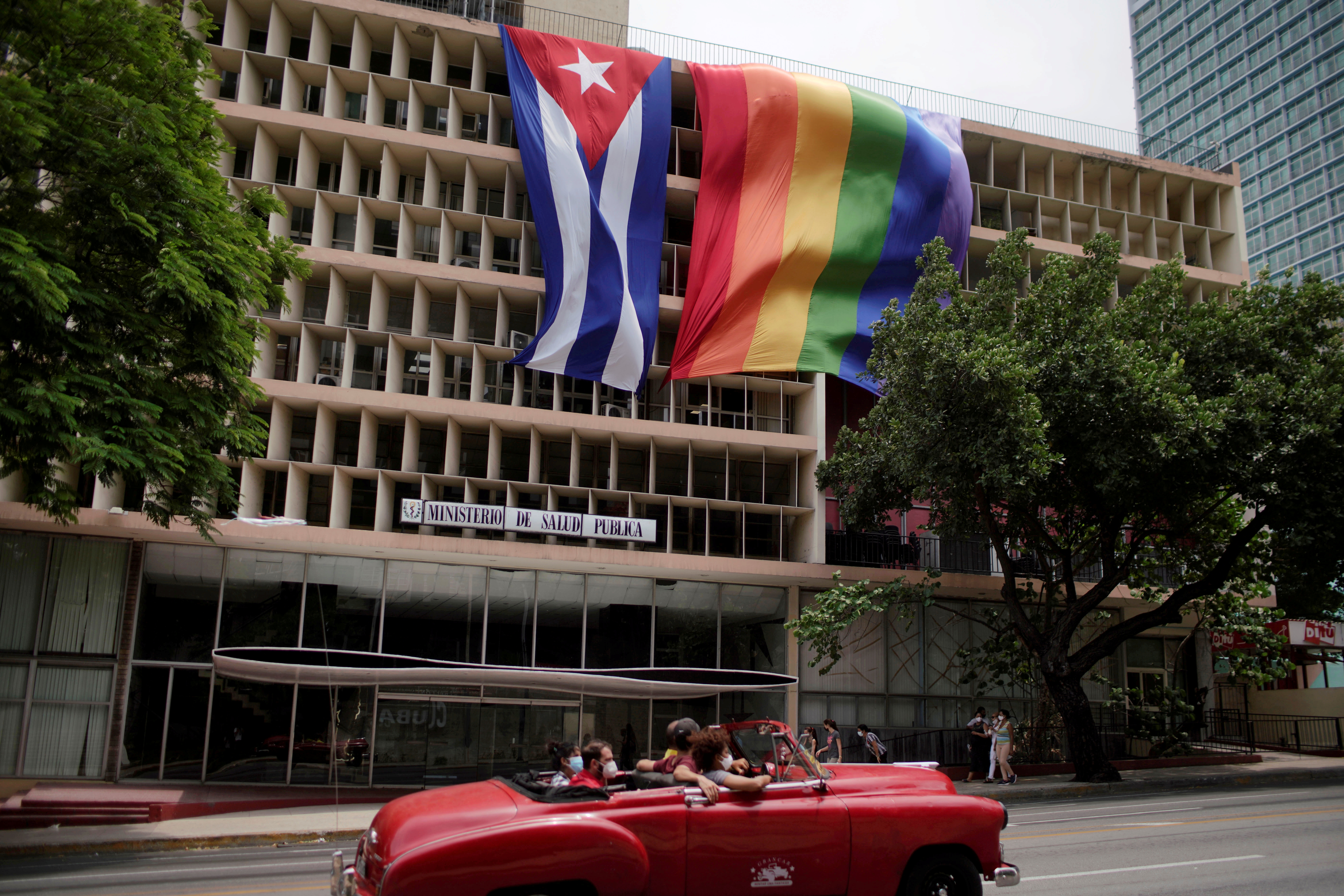 People pass in a vintage car in front of a rainbow flag hanging beside a Cuban flag at the Health Ministry building in Havana, Cuba, May 17, 2021. REUTERS/Alexandre Meneghini
