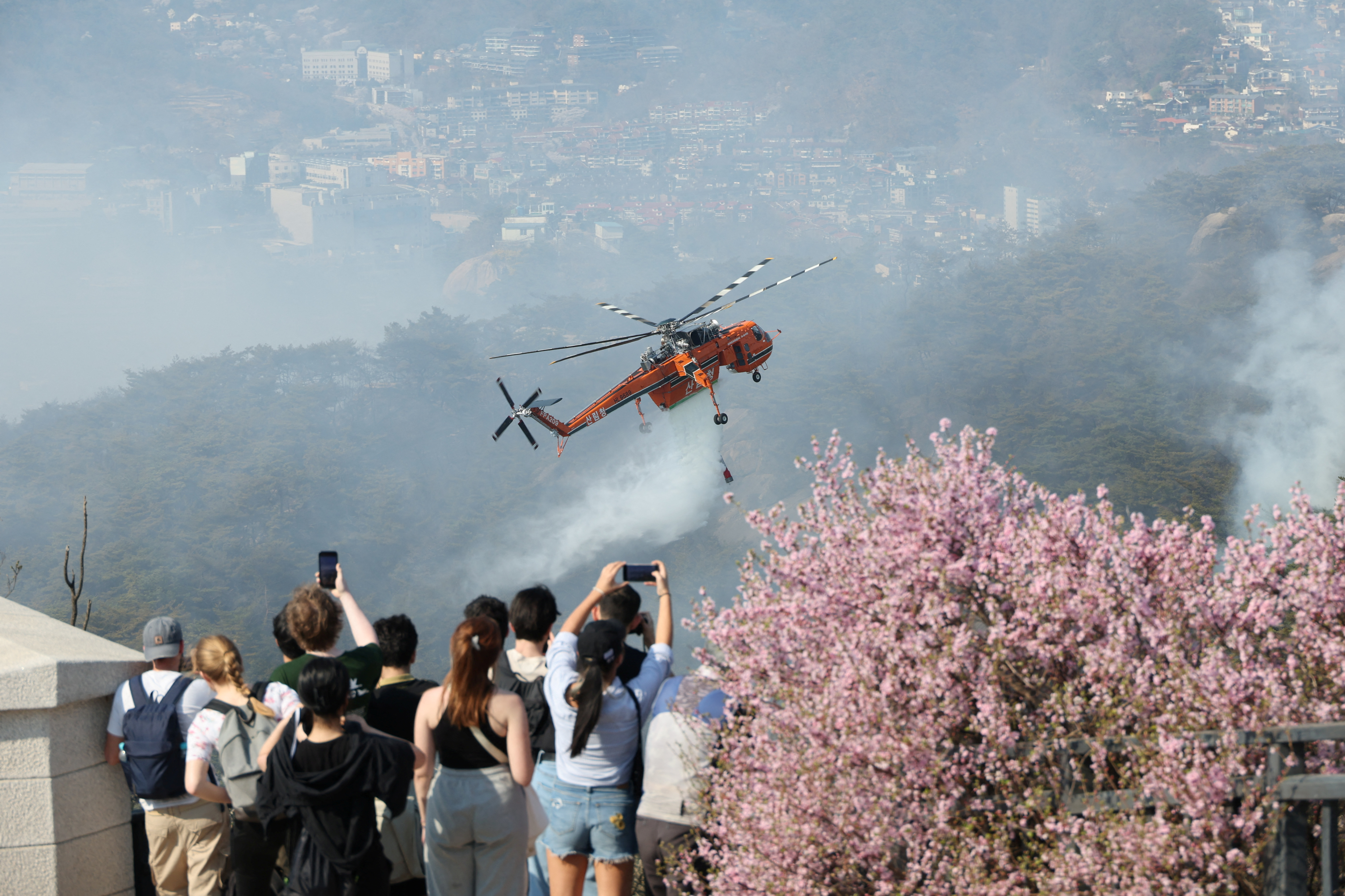 People watch a helicopter dropping water on fire at Inwangsan mountain in Seoul