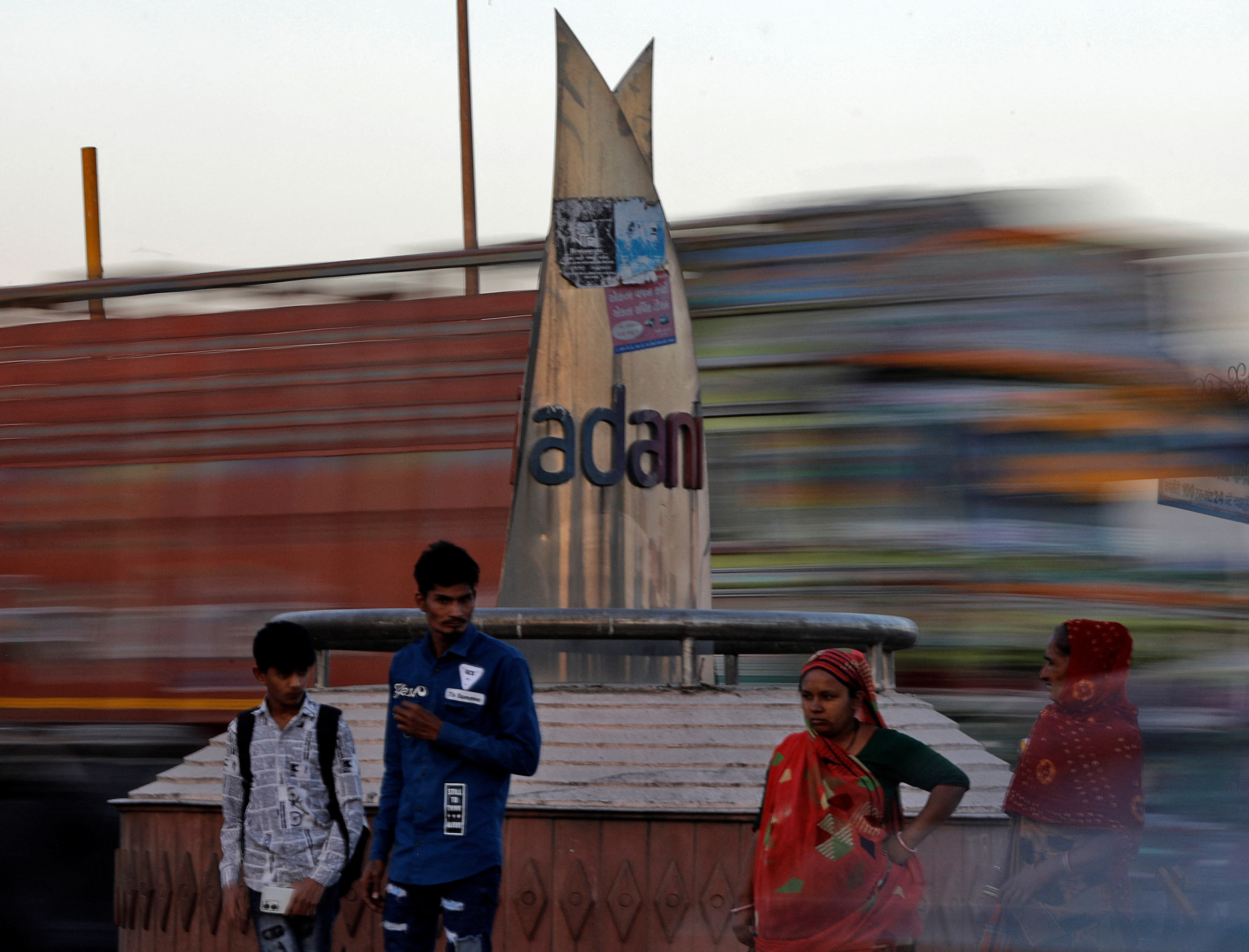 People wait to cross a road in front of the logo of the Adani Group installed at a roundabout on the ring road in Ahmedabad