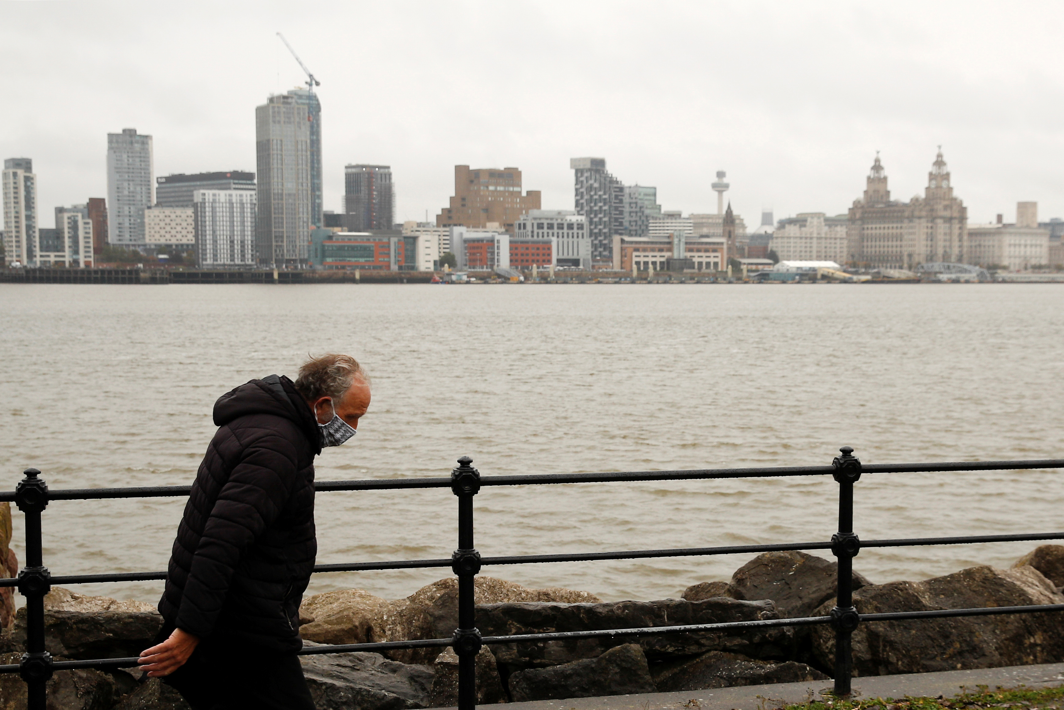 A man wearing a face mask walks along the River Mersey with the Liverpool skyline in Seacombe
