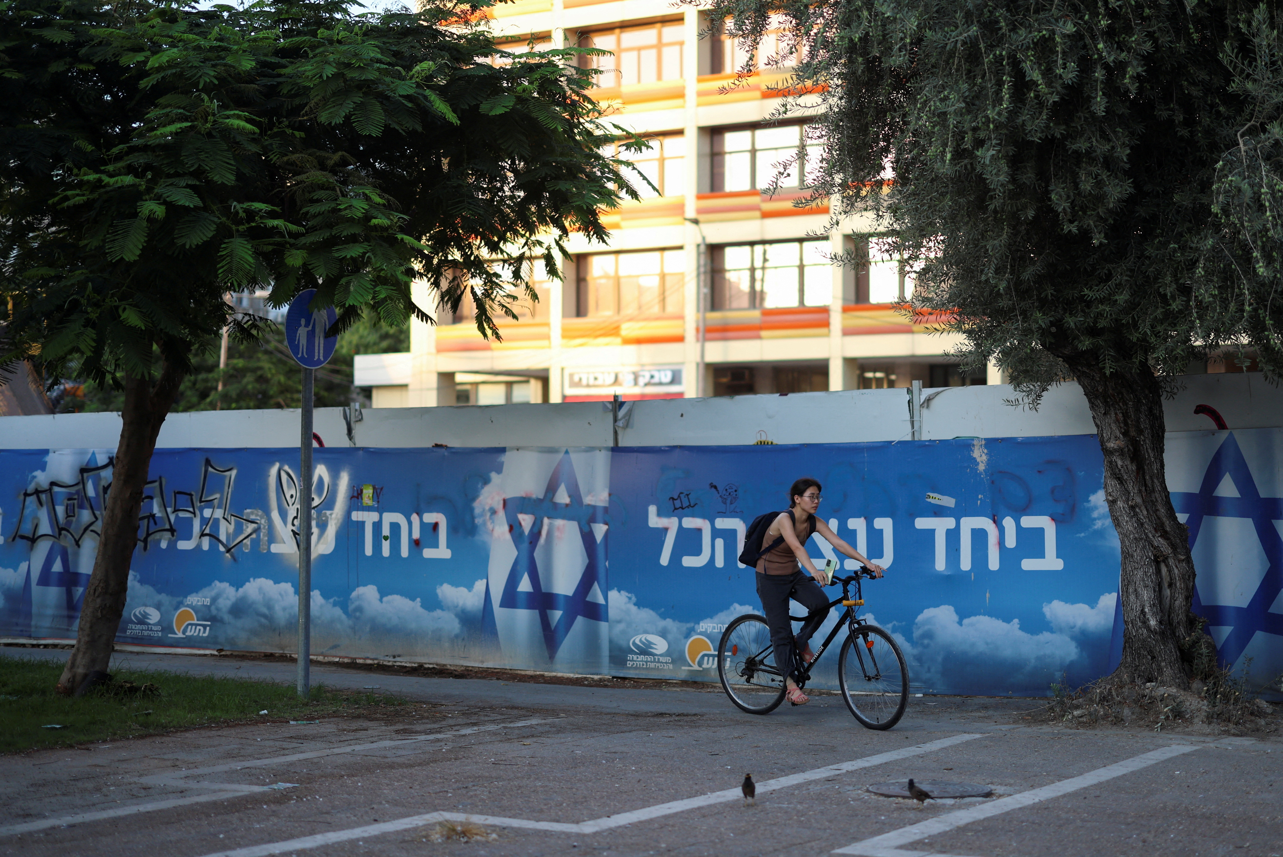 A woman on a bicycle rides past a billboard with the Israeli flag and a slogan in support of the hostages kidnapped during the deadly October 7 attack displayed, in Tel Aviv