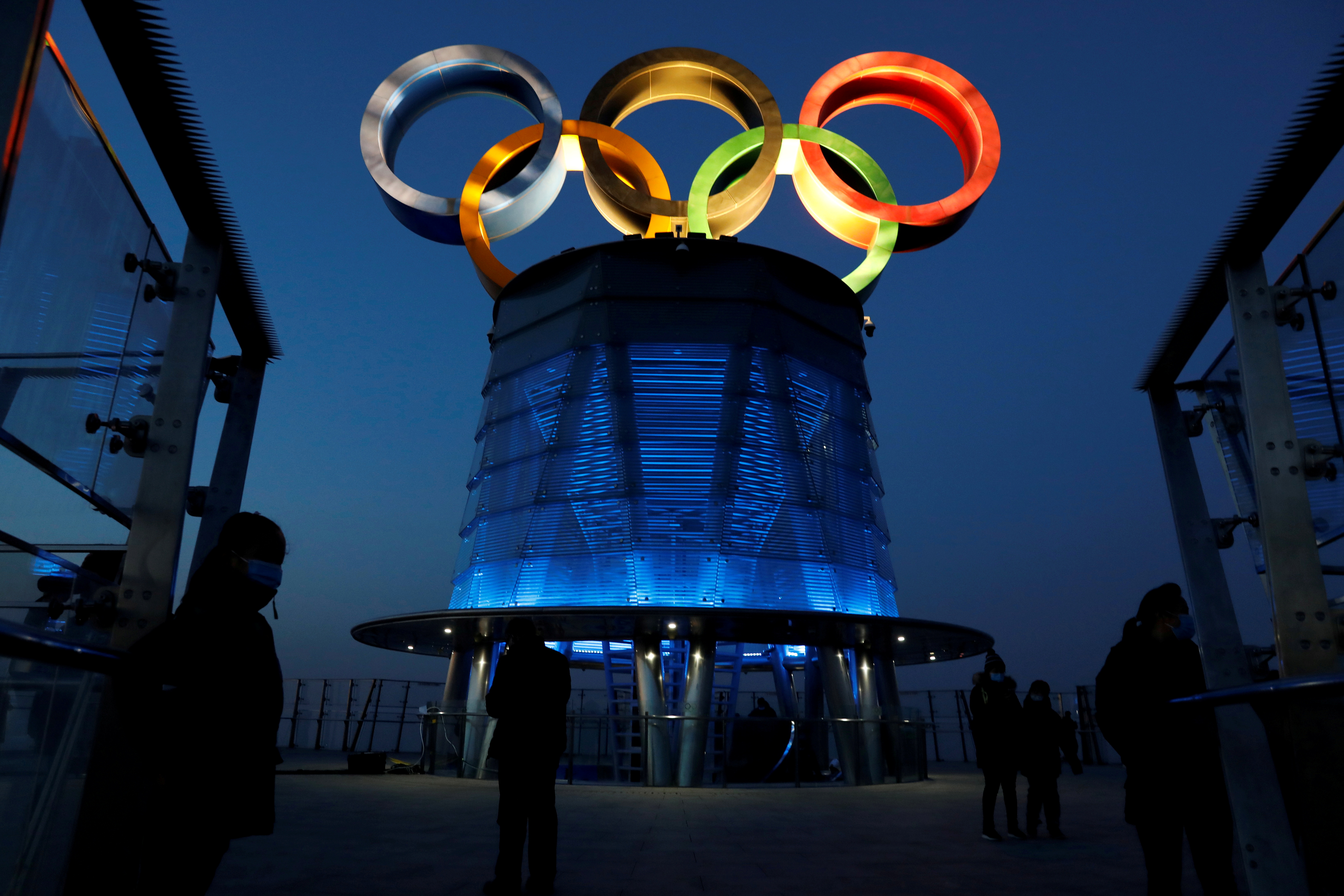 People wearing face masks following the coronavirus disease (COVID-19) outbreak are seen near the lit-up Olympic rings at top of the Olympic Tower, a year ahead of the opening of the 2022 Winter Olympic Games, in Beijing, China February 4, 2021. REUTERS/Tingshu Wang