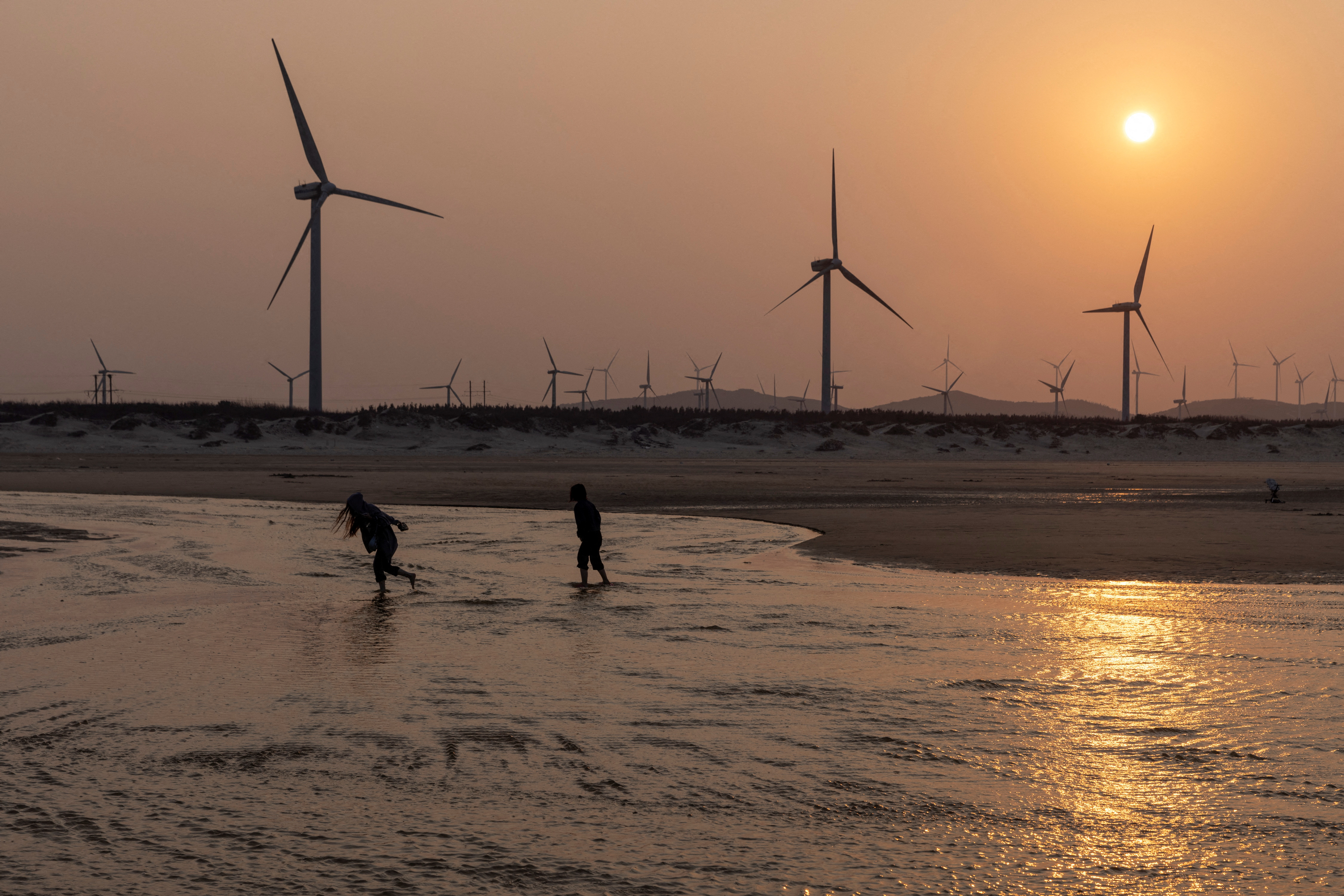 People walk past wind turbines at a beach on the Taiwan strait during sunset on Pingtan Island