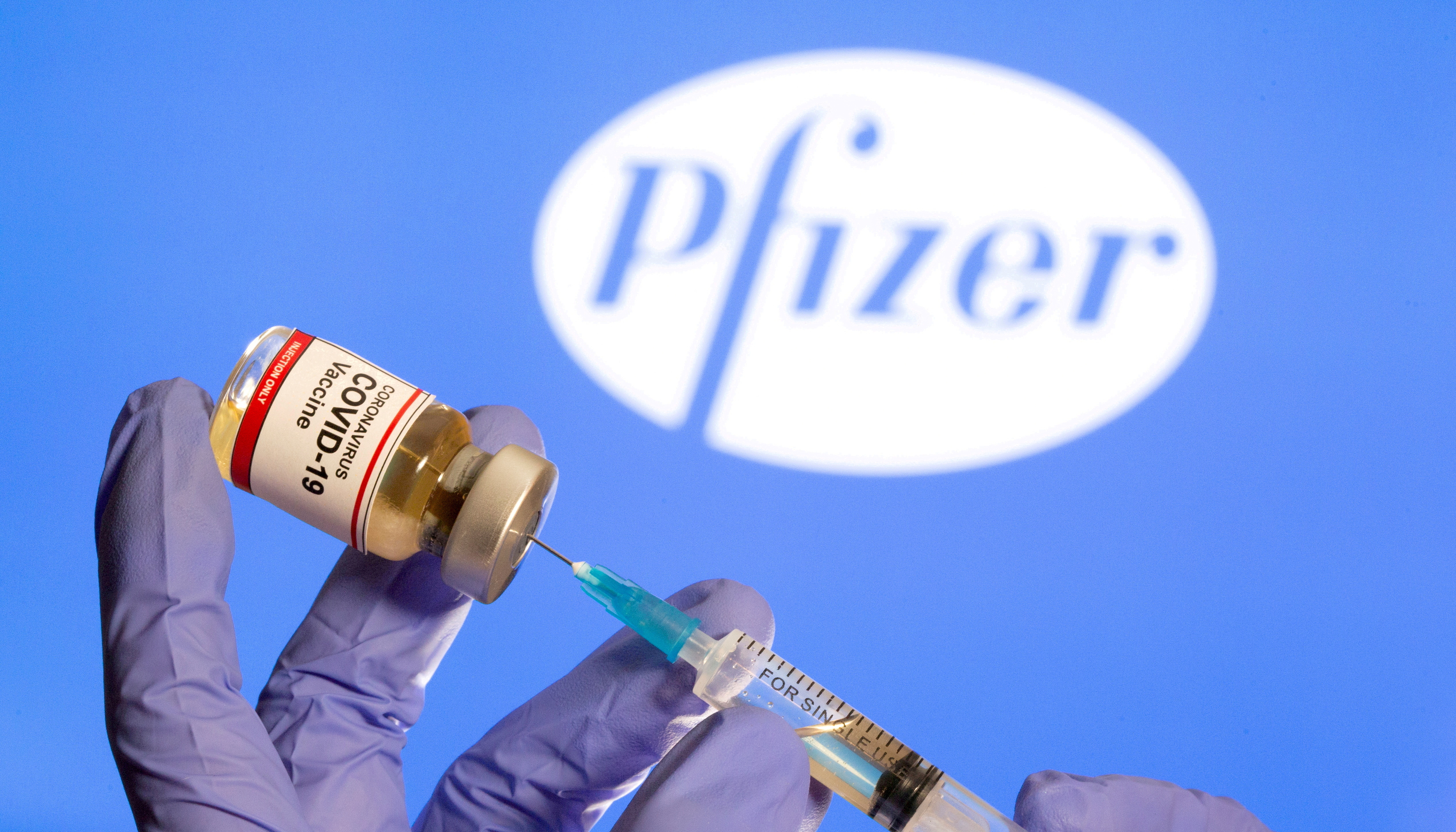 A woman holds a small bottle labeled with a 'Coronavirus COVID-19 Vaccine' sticker and a medical syringe in front of displayed Pfizer logo in this illustration